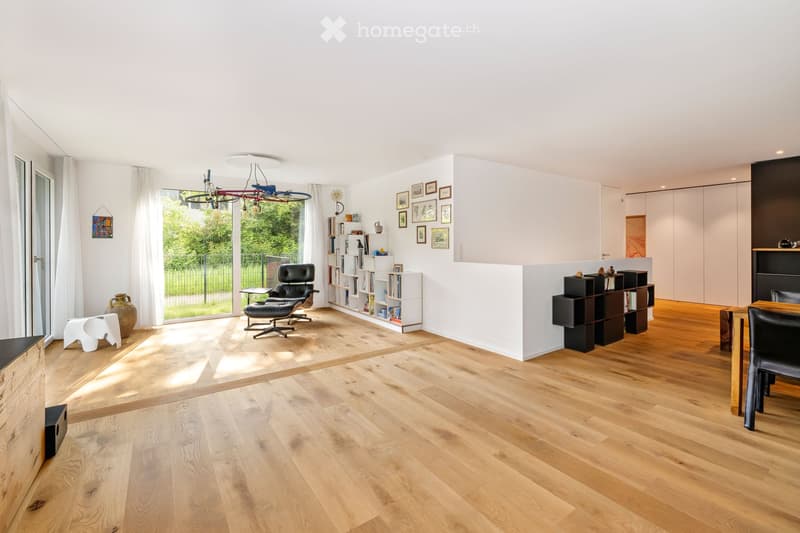 Bright living room with wooden flooring, large windows, modern furniture including a sofa, chair, table, and bookshelves.
