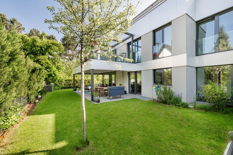 Modern house with a spacious garden, featuring a well-maintained lawn, patio area with outdoor furniture, and a barbecue. The house has large windows and a sleek, contemporary design.