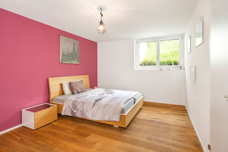 Modern bedroom with wooden flooring, a bed with a grey duvet and pillows, a wooden nightstand, a picture frame on the wall, and a pendant light fixture.