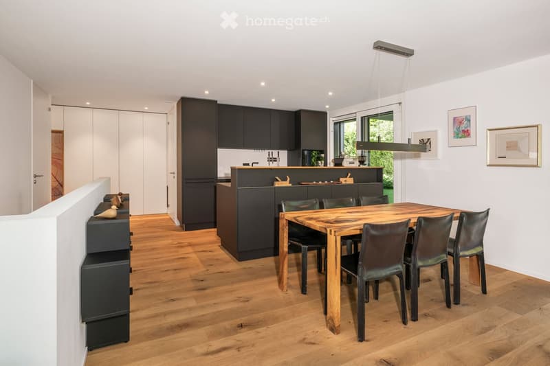 Modern kitchen with dark cabinets, a wooden dining table surrounded by black leather chairs, and a kitchen island with a countertop. The space features a pendant light fixture and framed artwork on the walls.