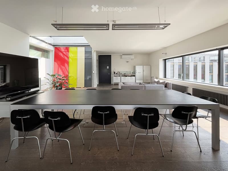 Modern, open-plan room with a large central table surrounded by chairs, a sofa area, a small kitchen area with a refrigerator, and a potted plant. Large windows and pendant lights are visible.