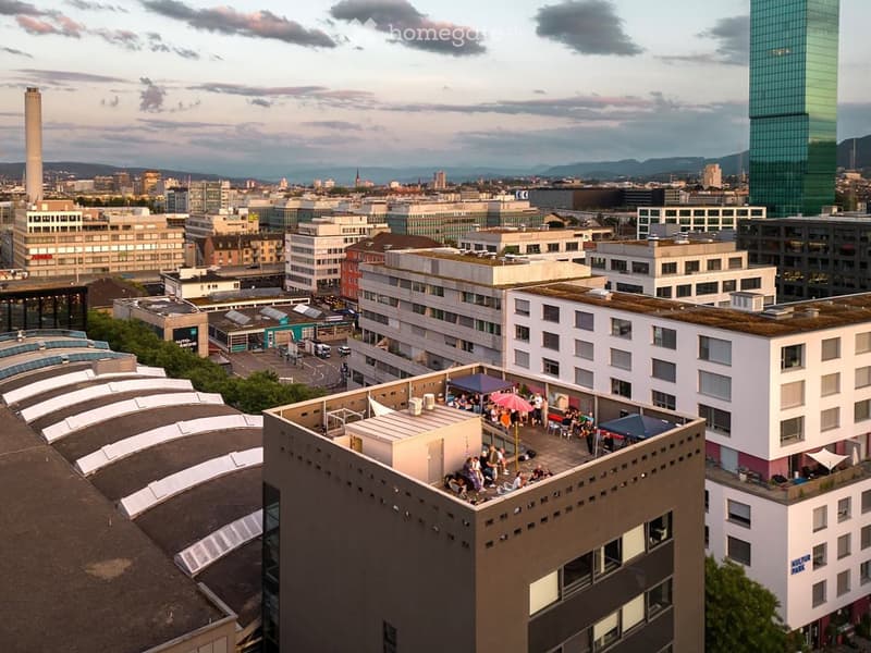 Aerial view of a cityscape with multiple buildings, including a rooftop terrace with people, umbrellas, and tables, surrounded by urban structures and distant mountains.