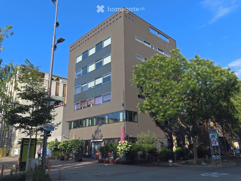 Modern multi-story building with large windows and a flat roof, surrounded by trees and greenery.