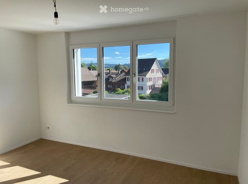 Empty room with wooden flooring, white walls, and large windows offering a view of neighboring houses and a clear blue sky.