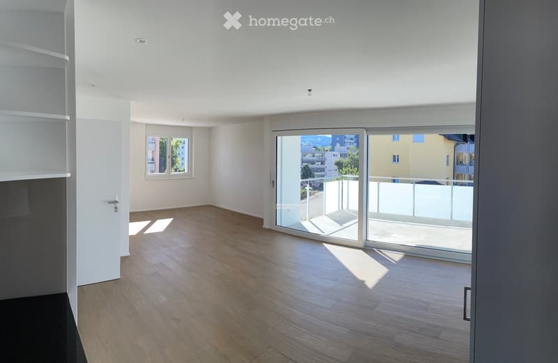 Empty room with wooden flooring, white walls, large sliding glass doors leading to a balcony, and a window on the opposite wall.