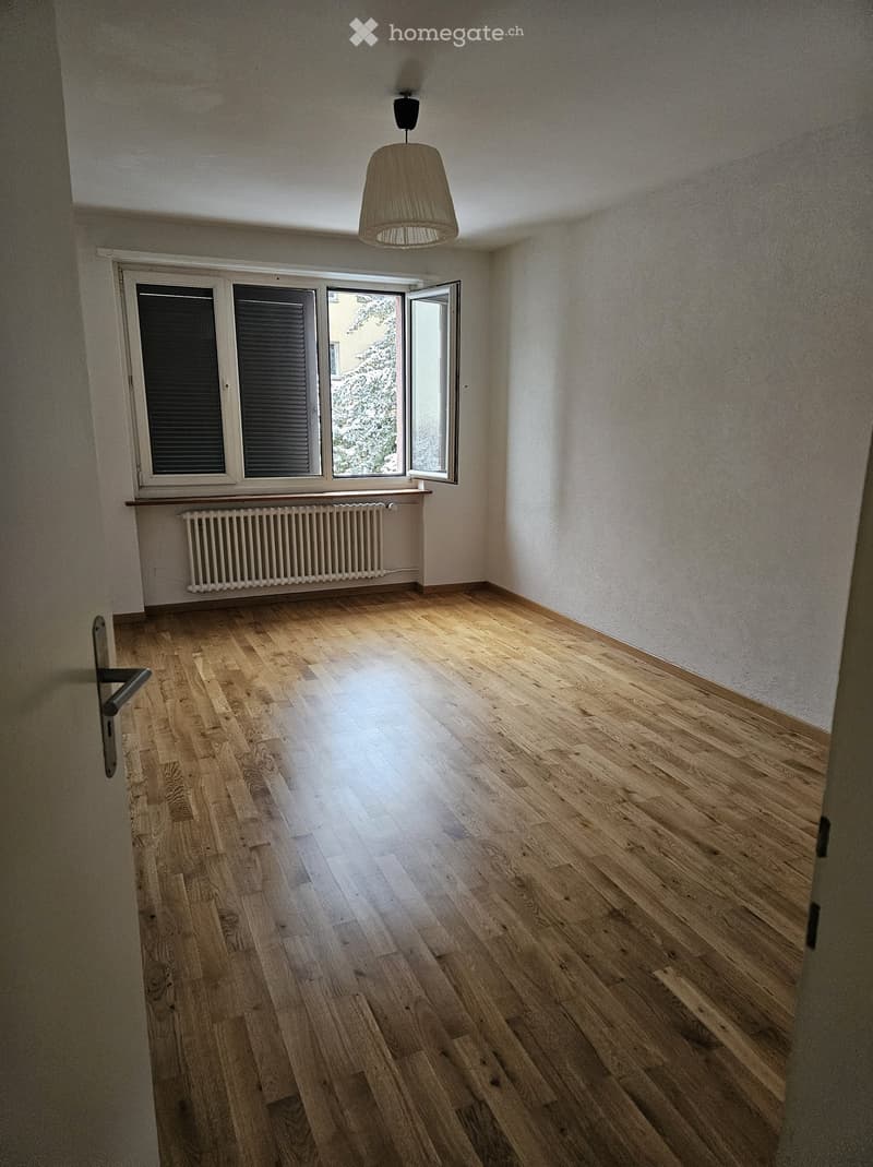 Empty room with wooden flooring, white walls, and a large window with blinds. A radiator is visible below the window, and a pendant light hangs from the ceiling.