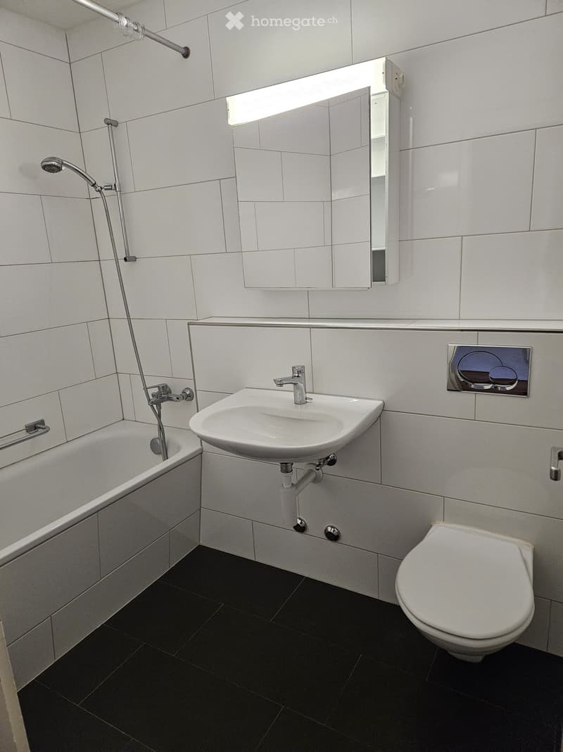 Modern bathroom with white tiles, a bathtub, a sink with a modern faucet, a toilet, a mirror, and a showerhead.