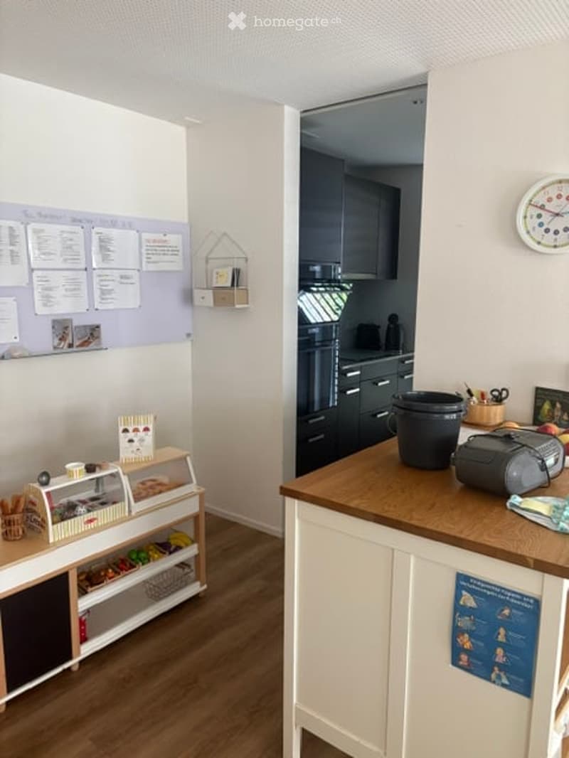A small, well-lit room with a display case, shelves, a clock, and a counter with a fridge and other items.