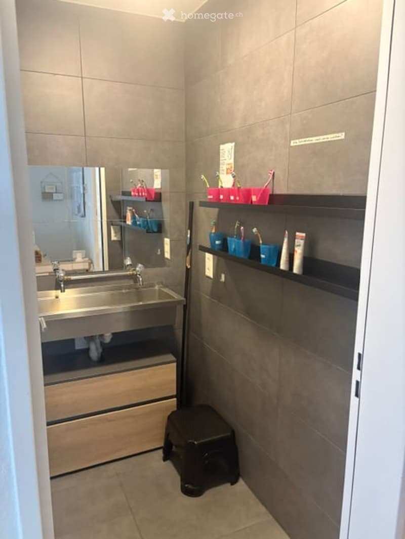 Modern bathroom with a double sink, large mirror, and built-in storage shelves holding toothbrushes and toothpaste. A small black stool is placed in front of the sink.