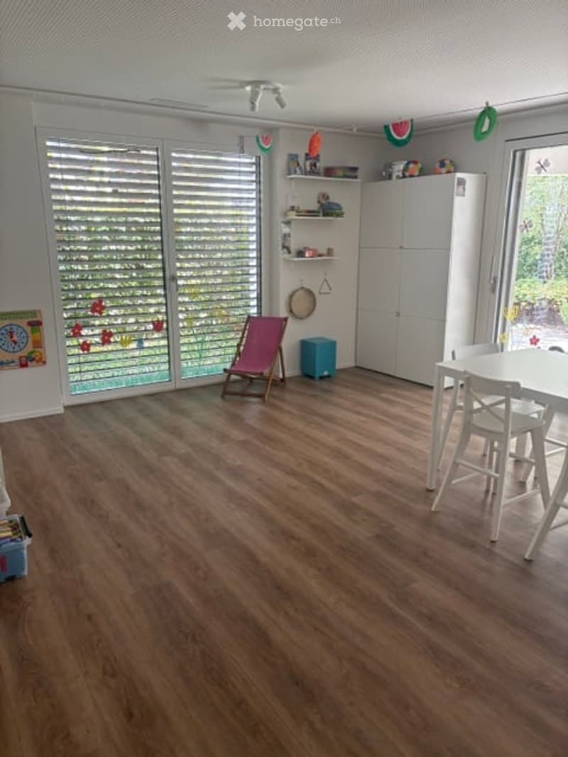 Bright room with wooden flooring, large windows with blinds, a pink folding chair, a white dining table with chairs, a blue storage box, and a white cabinet with shelves.