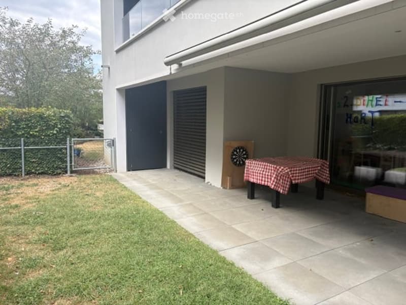 Outdoor patio area with a checkered tablecloth-covered table, dartboard, and a small seating area. The area is adjacent to a modern building with garage doors and a fenced yard.
