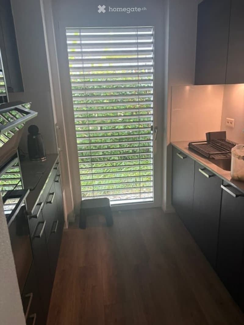 Modern kitchen with dark grey cabinets, wooden flooring, and a large window with blinds. Appliances include a stove, oven, fridge, and dishwasher. A small stool is placed near the window.