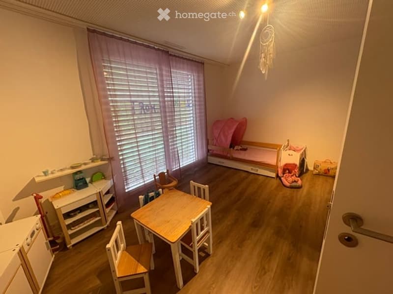 A well-lit children's room with a wooden floor, a small table and chairs, a toy bed, a toy crib, and a toy basket. The room features a large window with blinds and a pink curtain.