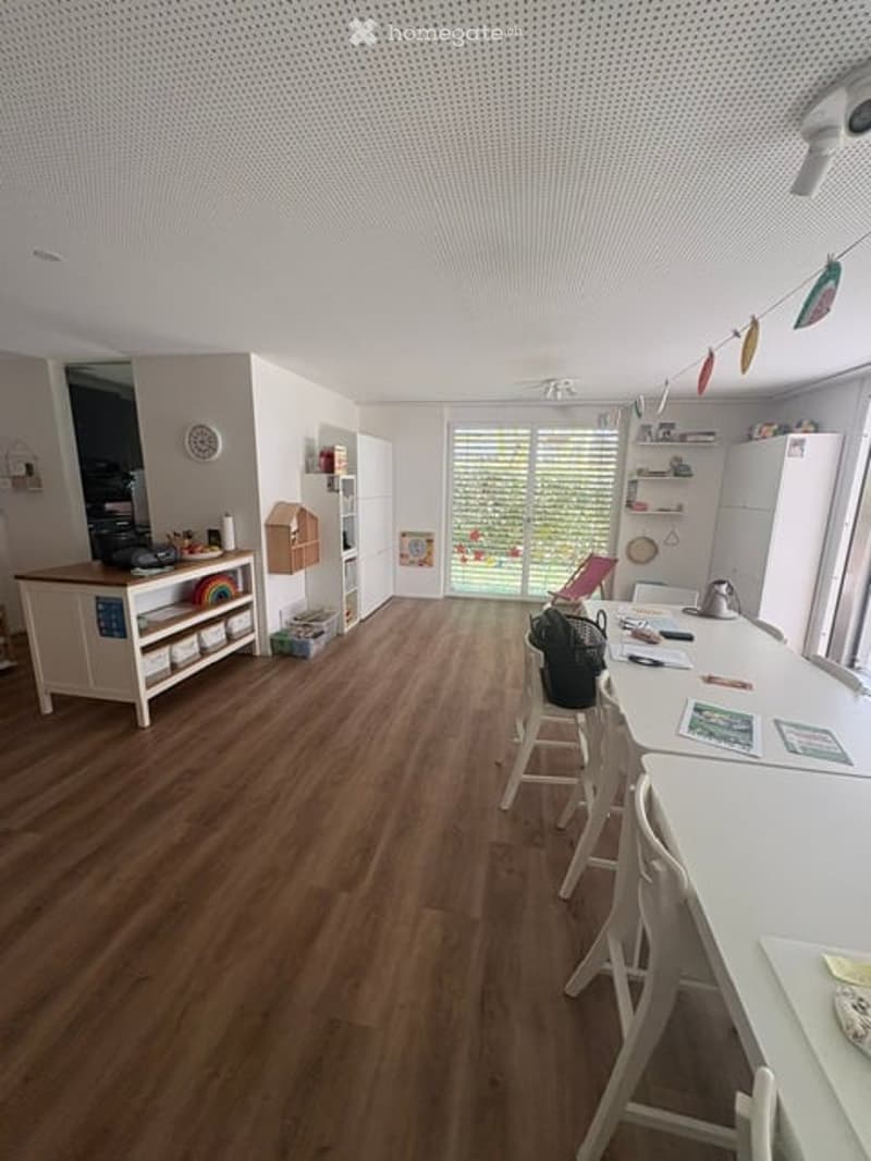 Bright, open kitchen with wooden flooring, white cabinets, and a large island with bar stools. A refrigerator, shelves, and a clock are visible.