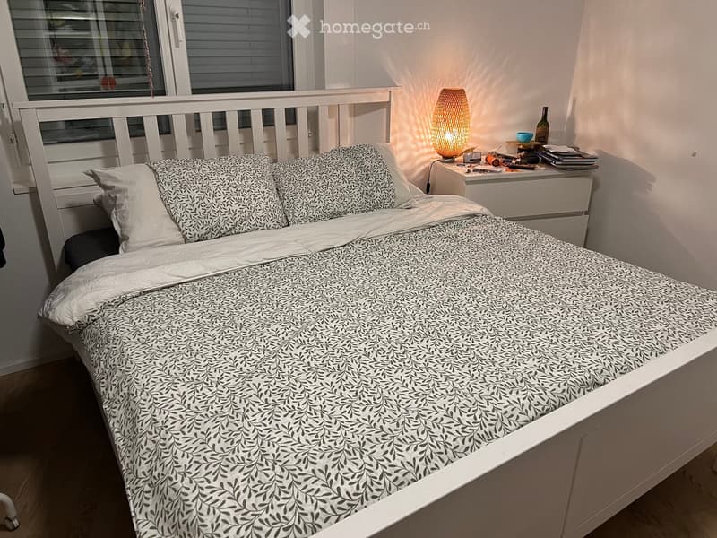 Cozy bedroom with a white bed, patterned bedding, a nightstand with a lamp, and a window with blinds.
