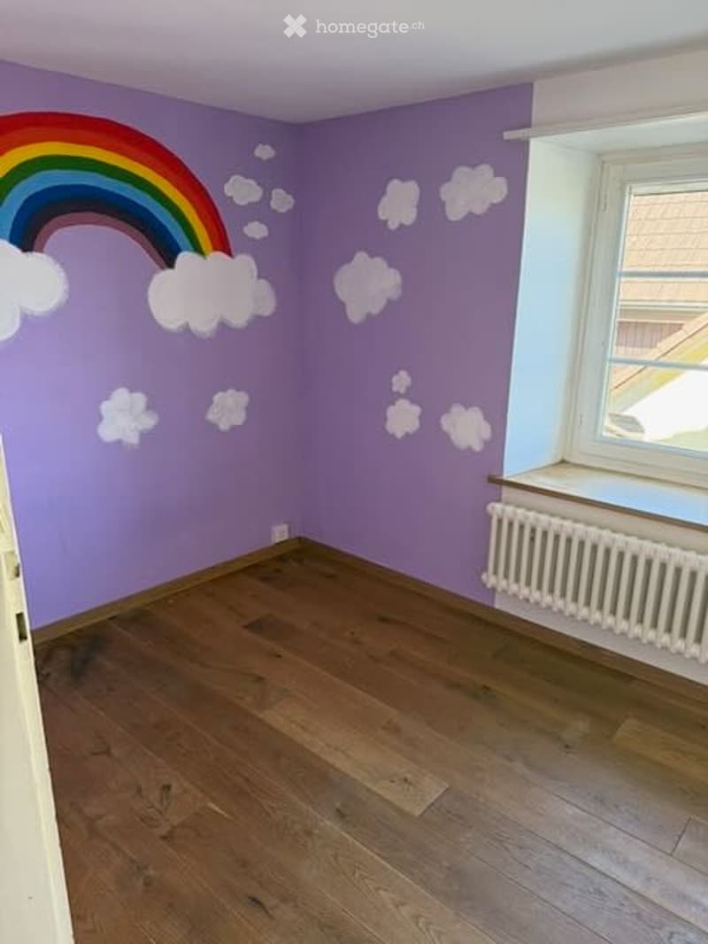 Empty room with wooden flooring and purple walls featuring a painted rainbow and cloud design. A white radiator is visible beneath a window.