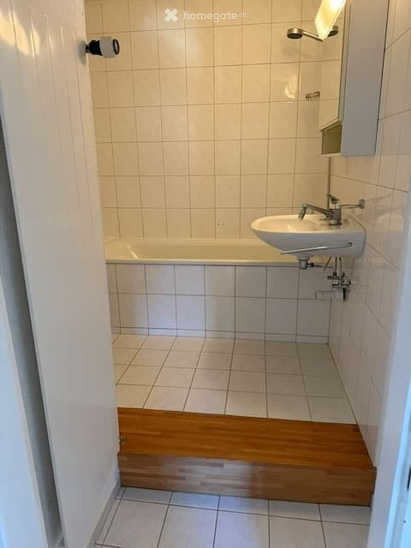 A small bathroom with a bathtub, sink, and mirror. The walls and floor are tiled in light beige, and there is a wooden step leading to the bathtub.