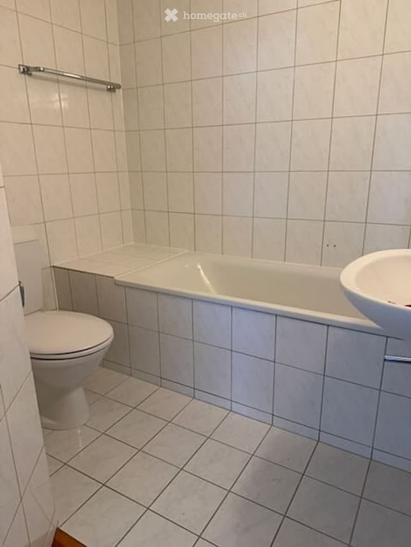 A clean, tiled bathroom with a bathtub, toilet, and sink. The walls and floor are covered in light gray tiles, and a towel rack is mounted on the wall above the bathtub.