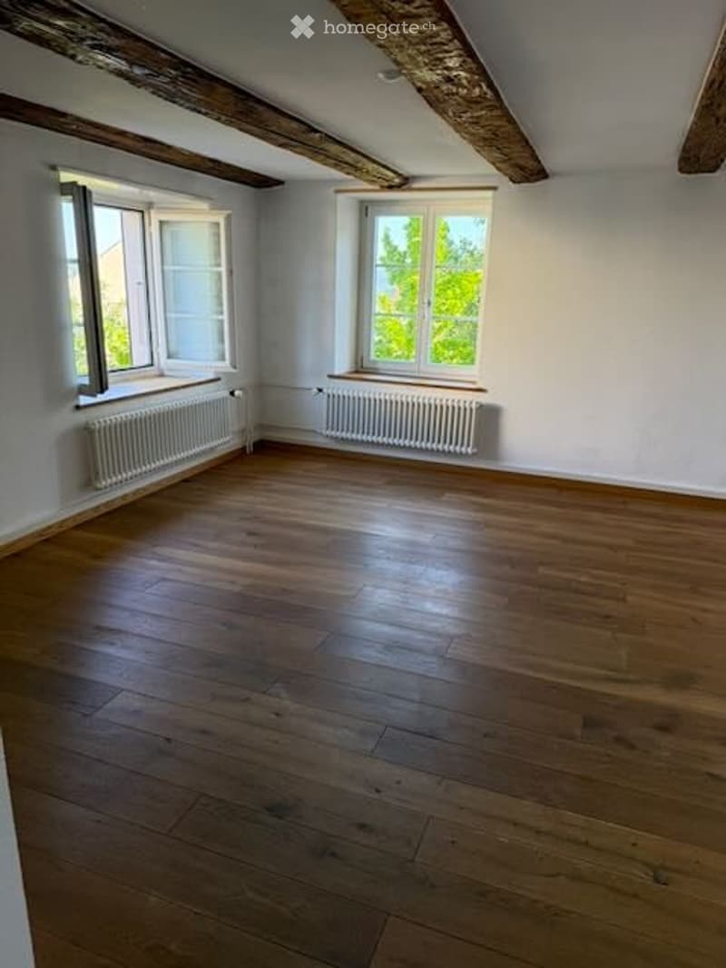 Empty room with wooden flooring, white walls, exposed wooden beams on the ceiling, and two windows with radiators beneath them.