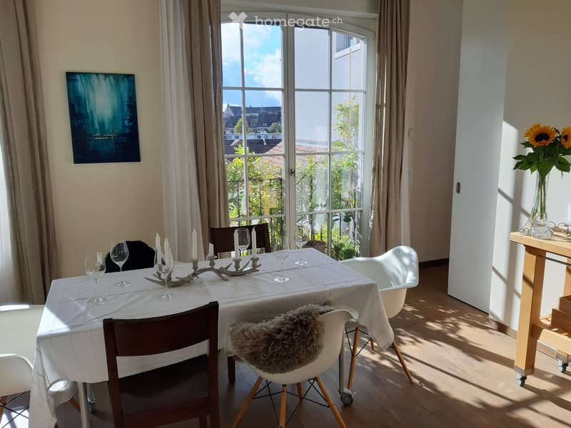 Bright dining area with a white tablecloth-covered table, surrounded by chairs, candle holders, and wine glasses. A vase with sunflowers sits on a small table near the window, which offers a view of the city and greenery outside.