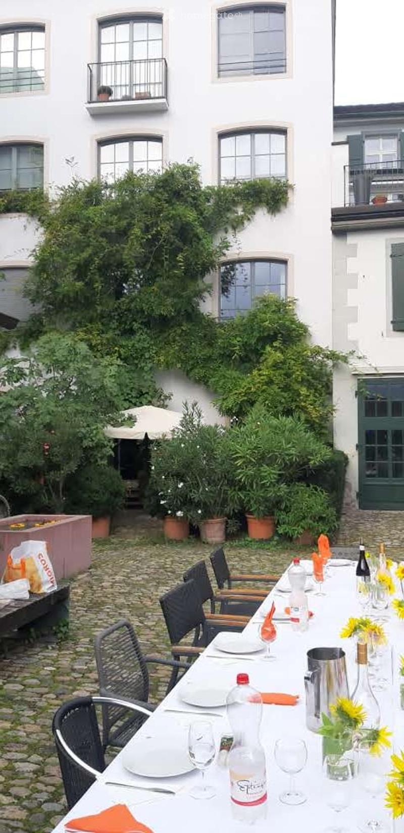 Courtyard with a long dining table set for a meal, surrounded by greenery and potted plants, with a building featuring balconies in the background.
