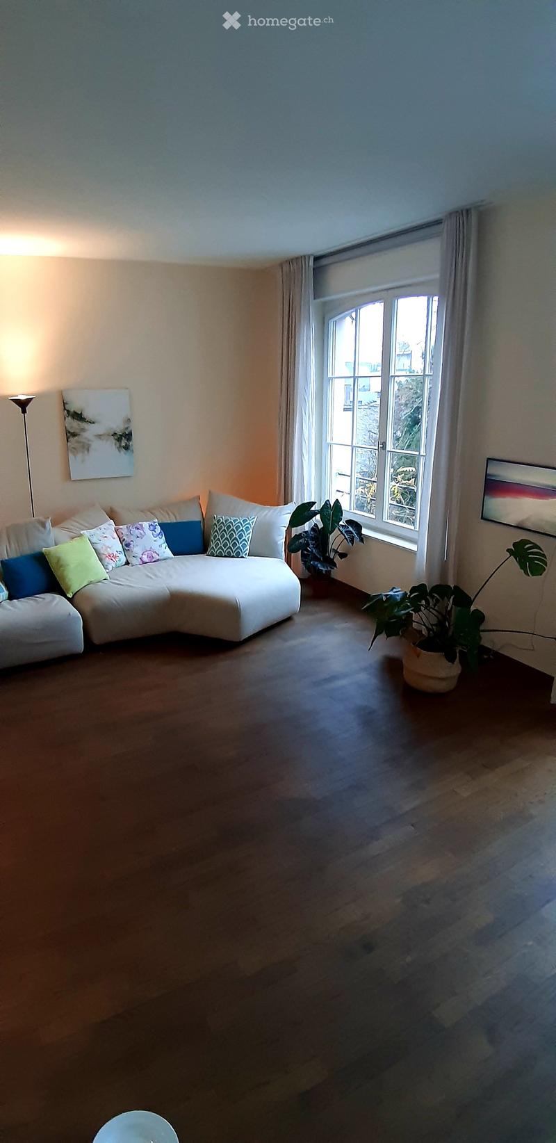 Modern living room with a white sectional sofa, colorful throw pillows, wooden floor, large window with curtains, and potted plants.
