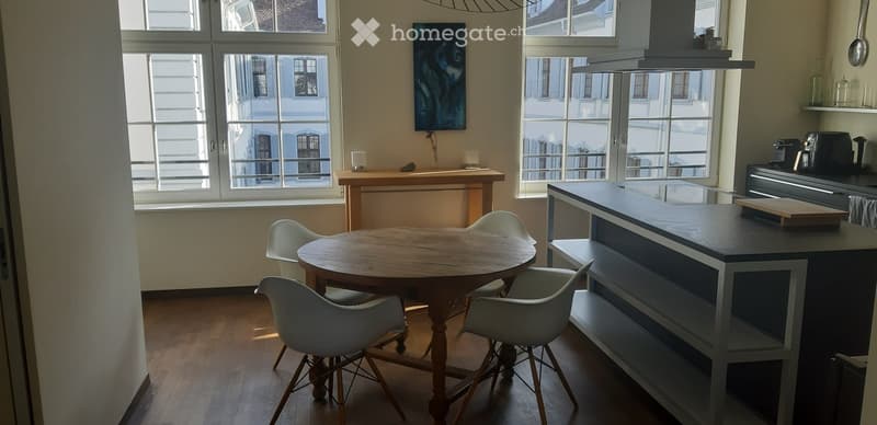 Bright, open-plan room with a wooden dining table and four white chairs, a modern kitchen island with a black countertop, and various appliances including a fridge, coffee maker, and teapot. Large windows offer a view of the city.