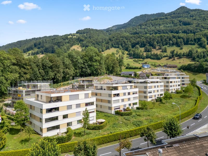 Aerial view of a modern residential complex with multiple buildings, surrounded by greenery and hills in the background.