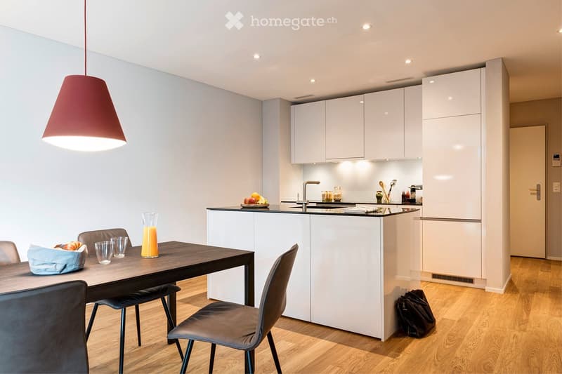 Modern kitchen with white cabinets, a wooden dining table, and four chairs. The counter has a fruit bowl, glasses, and a glass of orange juice. A red pendant light hangs above the table.