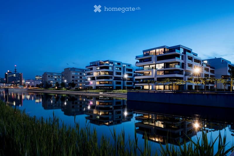 Modern multi-story residential buildings with illuminated windows and balconies, reflected in a calm water body at dusk.