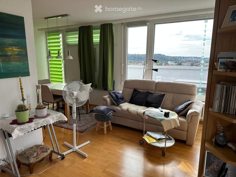 Modern living room with large windows offering a scenic view, featuring a beige sofa with dark blue pillows, a white fan, a small coffee table with a remote control, a small side table with a plant, and a dining area with a table and chairs.