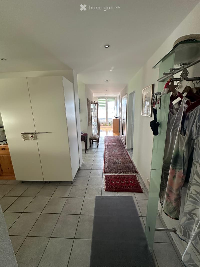 A well-lit hallway with tiled flooring, multiple rugs, and a coat rack with hanging items. The hallway leads to an open area with large windows and a view of another room.