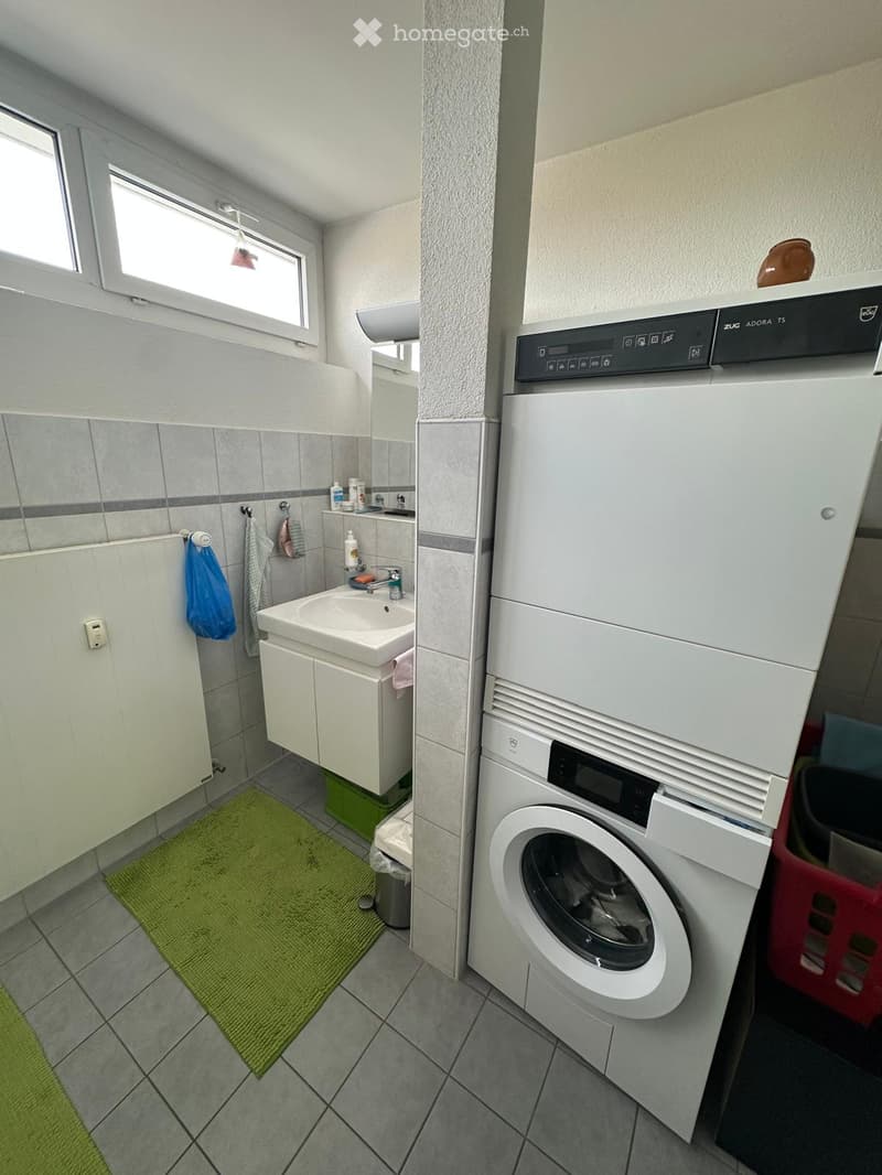 Bathroom with a sink, mirror, and a washing machine and dryer unit. The sink has a faucet and a small cabinet underneath. A green rug is on the floor, and a towel hangs on a rack.