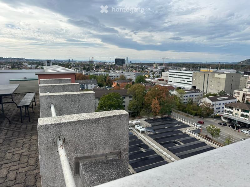 Roof terrace with a table and bench, overlooking a cityscape with buildings, trees, and a parking lot.