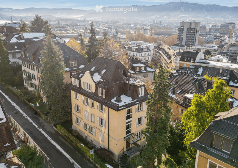 A multi-story residential building with a distinctive roof and white-framed windows, surrounded by other buildings and trees in a cityscape with distant mountains.