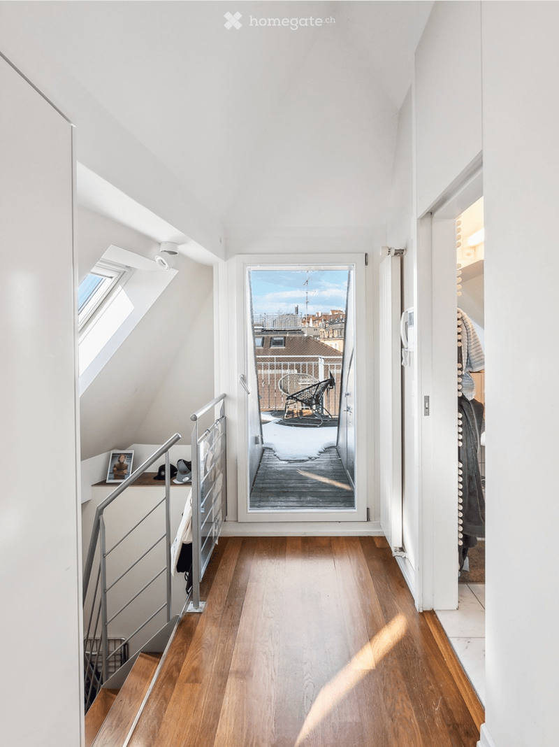 Narrow hallway with wooden flooring, white walls, and a metal railing leading to a small balcony area with outdoor furniture.