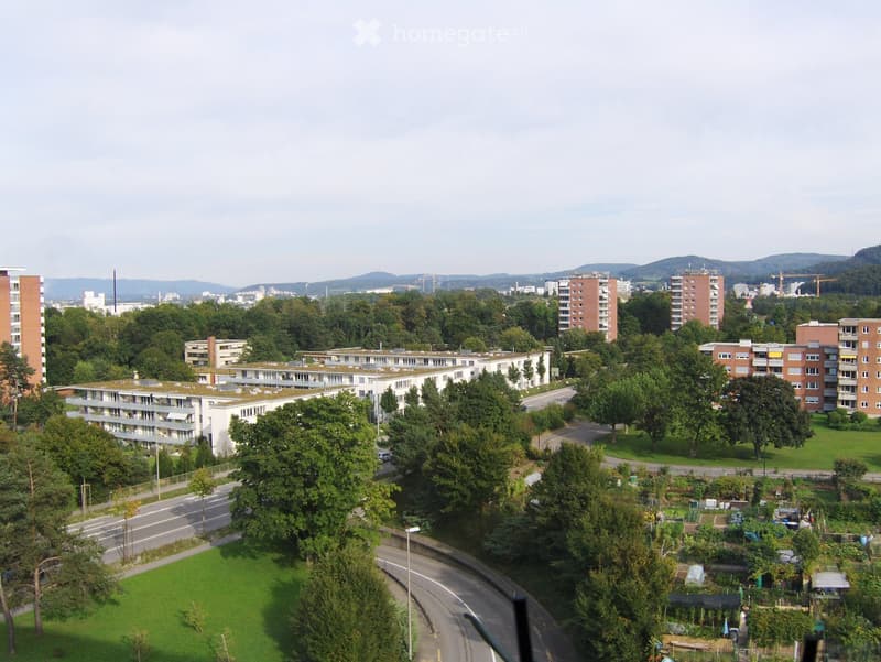 Aerial view of a residential area with multiple apartment buildings, green spaces, and trees, surrounded by a road network and distant hills.