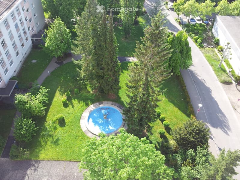 Aerial view of a well-maintained garden area with a central swimming pool, surrounded by trees, shrubs, and pathways. The garden is adjacent to a multi-story building with balconies and windows.