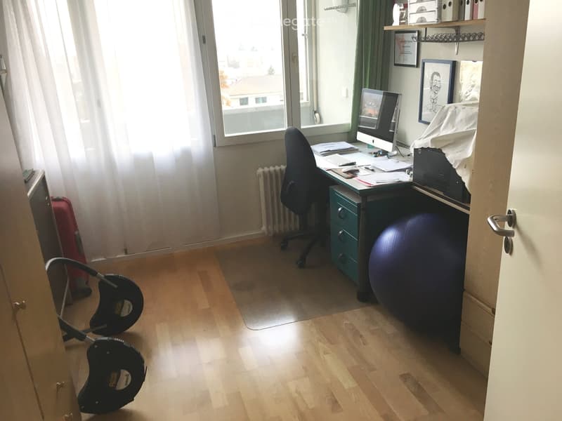A well-lit room with wooden flooring, a desk with a computer, a chair, a large exercise ball, and framed pictures on the wall. A window with curtains offers a view of the outside.