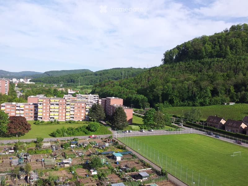 Aerial view of a residential area with apartment buildings, green spaces, a soccer field, and surrounding hills.