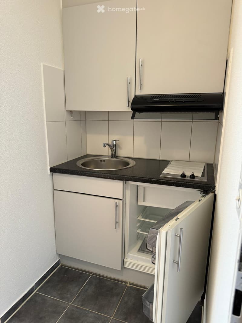 Compact kitchen with white cabinets, black countertop, stainless steel sink, and small refrigerator.
