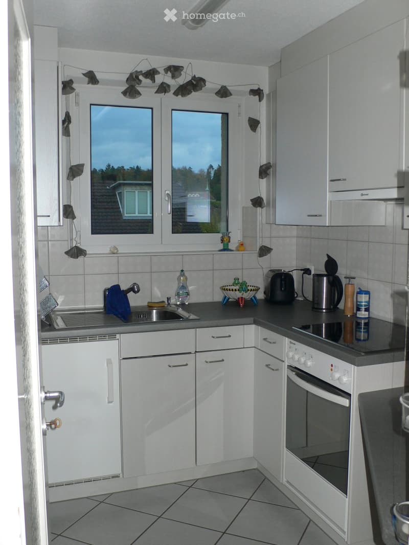Modern kitchen with white cabinets, grey countertops, and tiled flooring. Features a sink, oven, toaster, coffee maker, and various small appliances and decorative items.