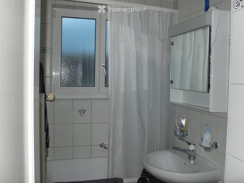A small bathroom with a white sink, mirror cabinet, and shower area with a curtain. The walls are tiled, and a window is visible above the shower.