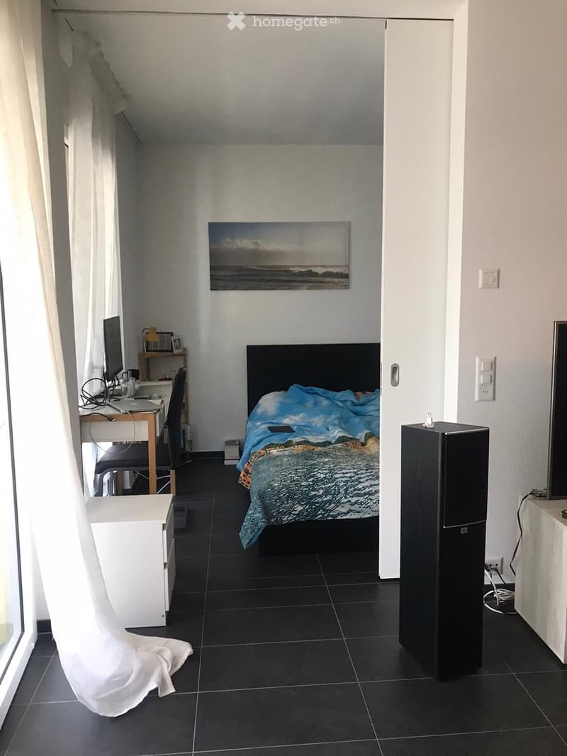 A modern bedroom with a bed, desk, chair, and a painting on the wall. The room features dark tiled flooring and white walls.