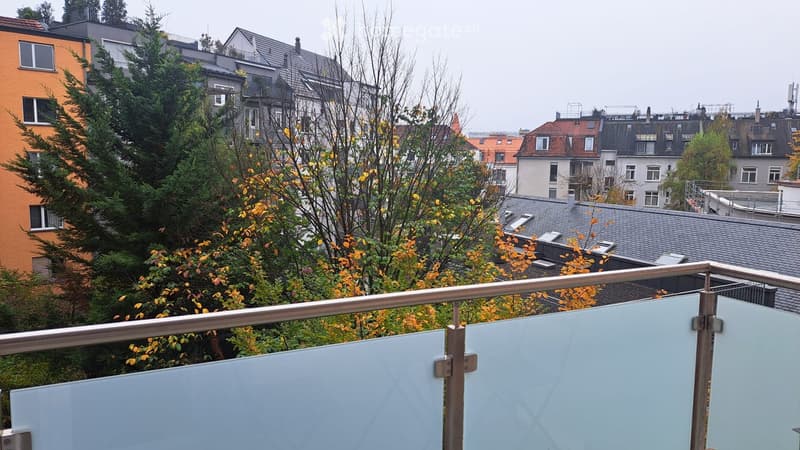 Balcony with metal railing and glass panels, overlooking a cityscape with multiple buildings, trees, and rooftops.