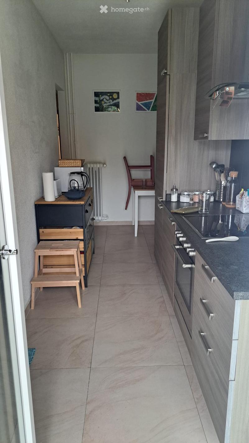 A narrow hallway with a modern kitchen on the right side, featuring dark grey cabinets, a black countertop, and various kitchen appliances. On the left, there is a small wooden table with a kettle, paper towels, and other items, along with a small wooden