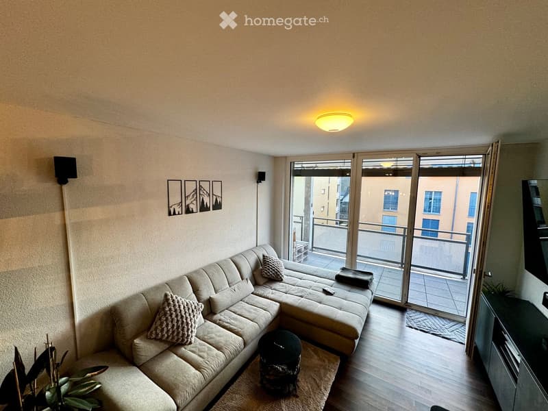 Modern living room with a large sectional sofa, coffee table, wall-mounted speakers, picture frames, and a plant near the entrance to a balcony.