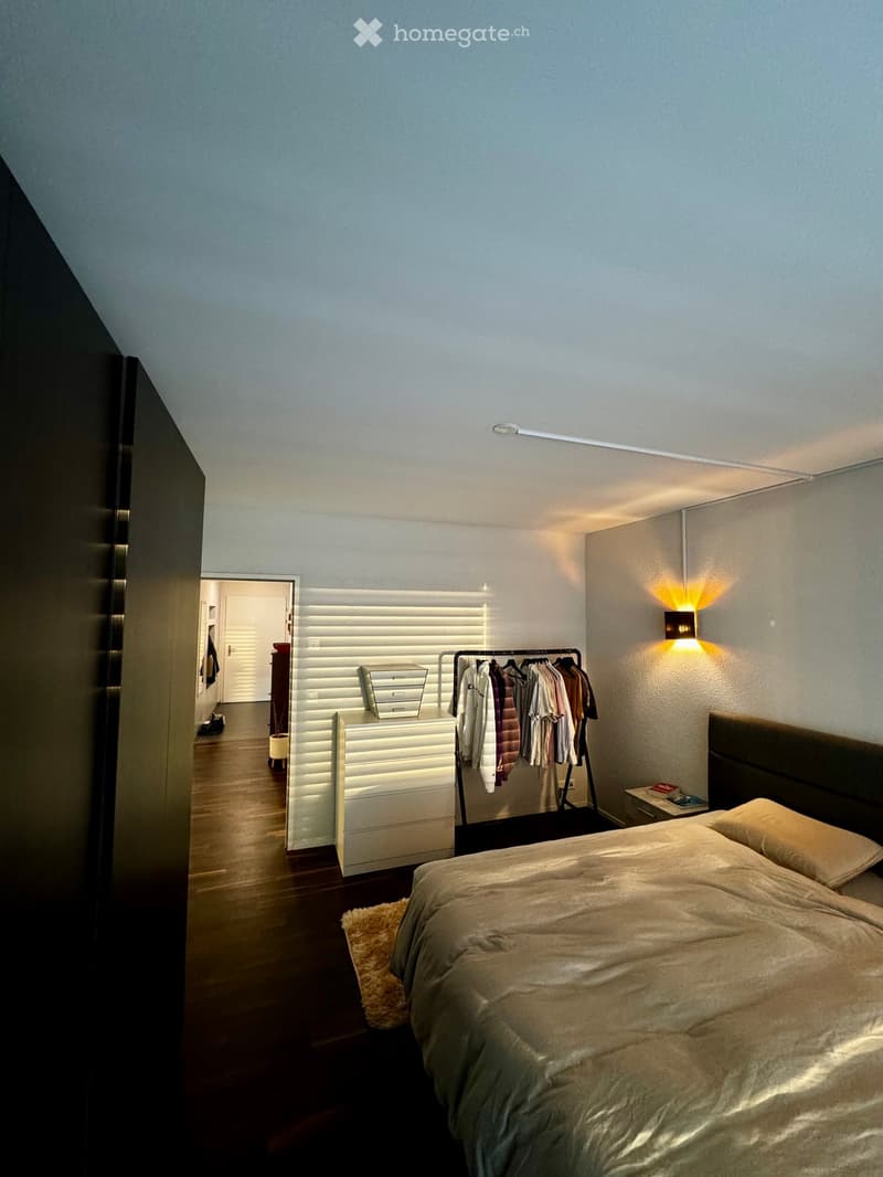 Modern bedroom with a large bed, nightstand, clothing rack, and a drawer unit. The room features wooden flooring and soft lighting.