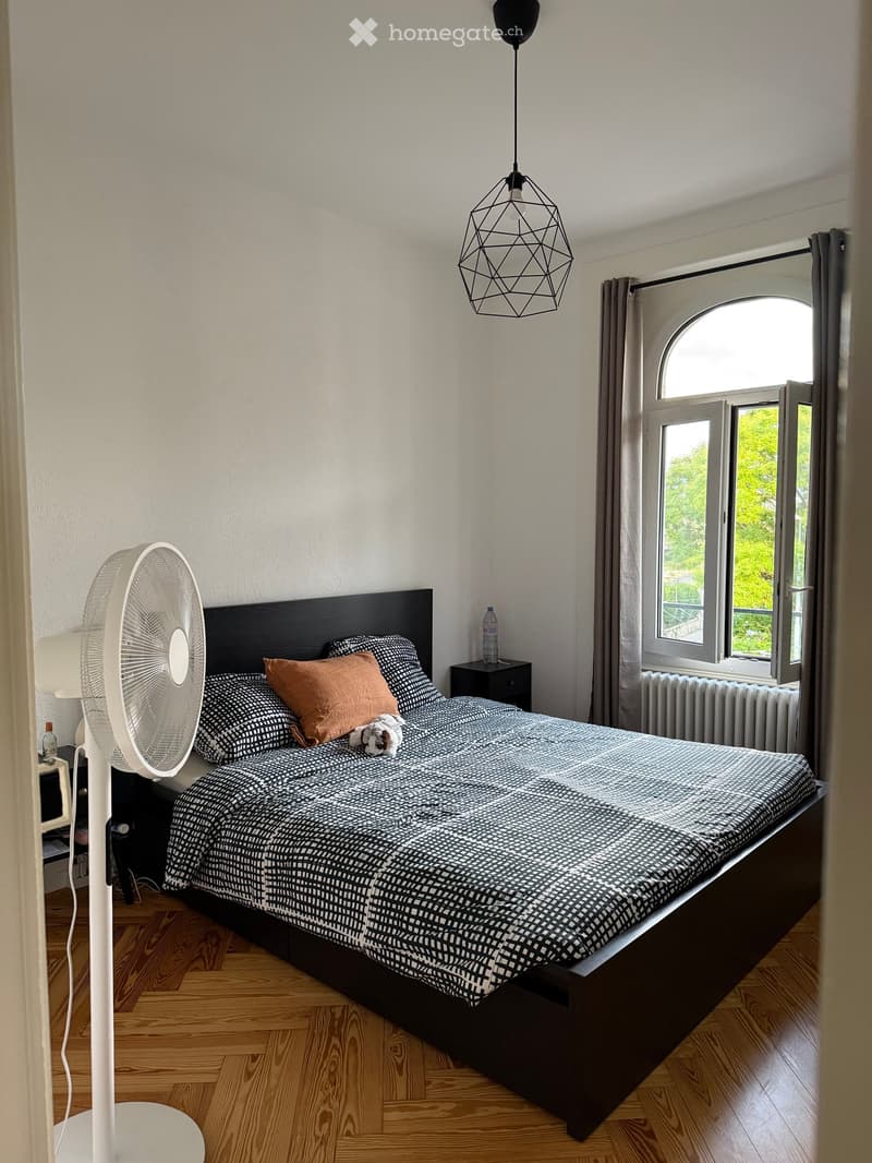 A modern bedroom with a wooden floor, a large bed with a black and white patterned duvet and pillows, a standing fan, a bedside table with a lamp, and a window with curtains.