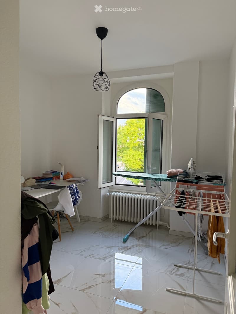 Bright room with white tiled floor, large arched window, desk with laptop and books, chair, ironing board, clothes hanger, radiator, and a modern hanging lamp.